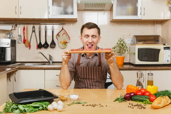 Молодой весельчак в фартуке сидит за столом с кусками мяса на деревянной доске, овощами, готовит дома мясной кол из свинины, говядины или баранины, на светлой кухне, полной посуды . — стоковое фото