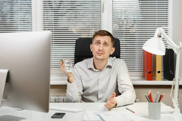 Impresionado y perplejo hombre de negocios en camisa sentado en el escritorio con mucho dinero en efectivo, trabajando en la computadora con monitor moderno, lámpara, documentos en la oficina de luz sobre el fondo de la ventana. Gerente o trabajador — Foto de Stock