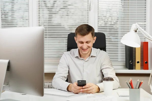 Hombre de negocios guapo sonriente en camisa sentado en el escritorio con teléfono móvil, trabajando en la computadora con monitor moderno, lámpara, carpetas, café o té, documentos en la oficina de luz en el fondo de la ventana . — Foto de Stock