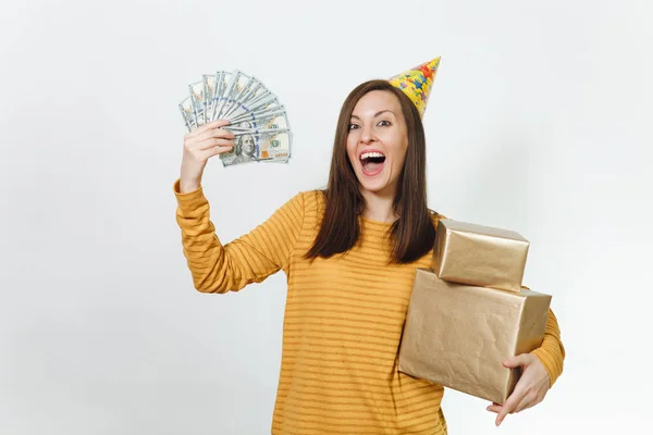 Caucasian fun young happy woman in yellow clothes, birthday party hat holding golden gift boxes with present and wad of cash money, celebrating holiday on white background isolated for advertisement.
