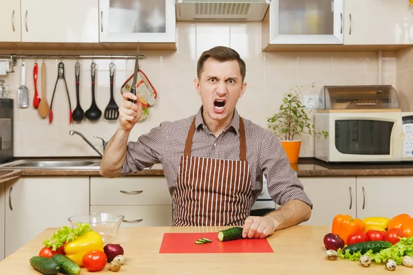 Zły szalony kaukaski młody człowiek w fartuch, brązowy shirt siedząc przy stole, atak kawałki warzyw na sałatkę z nożem w kuchni światło. Pojęcie diety. Zdrowy styl życia. Gotowanie w domu. Przygotowanie żywności. — Zdjęcie stockowe