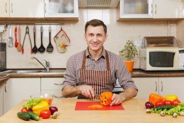 Gülümseyen beyaz yakışıklı genç apronda, kahverengi gömlek masadan, sebze salatası için ışık mutfakta bıçakla keser. Diyet kavramı. Sağlıklı bir yaşam tarzı. Evde yemek. Yemek hazırlamak.