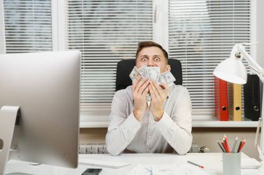 Çalışma ile modern sınıf başkanı bilgisayar, pencere arka plan üzerinde ışık office belgelerinde konusunda mutlu gülümseyen iş tişörtlü adam çok nakit para ile masaya oturuyor. Yöneticisi veya işçi kazanan.