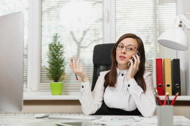 Işık Office belgeleri ile çağdaş bilgisayarda çalışan güzel ciddi iş kadın takım elbise ve masa başında oturan gözlük cep telefonu üzerinde sorunları çözme bir kenara seyir konuşurken
