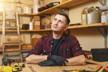 Yakışıklı beyaz genç adam ekose gömlek, siyah T-shirt, gülümseyen ahşap masa doğrama atölyesinde çalışan eldiven ile ahşap, farklı araçlar parça yer.