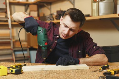 Yakışıklı gülümseyen beyaz genç adam ekose gömlek, siyah T-shirt, matkap parça ahşap, ahşap masa doğrama atölyesinde çalışan ile sondaj eldiven farklı araçlarla yerleştirin..