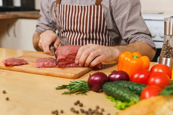 Εσωτερικη Καυκάσιος νεαρός άνδρας στην ποδιά που κάθεται στο τραπέζι με τα λαχανικά, το μαγείρεμα στο σπίτι προετοιμασία ποντάρισμα κρέατος από χοιρινό, βοδινό ή αρνί, ελαφριά κουζίνα με ξύλινη επιφάνεια, γεμάτη φανταχτερά μαγειρικά σκεύη. — Φωτογραφία Αρχείου