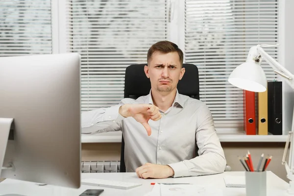 Serious tired and stress business man in shirt sitting at the desk, showing thumb down, working at computer with modern monitor, documents in light office on window background. Manager or worker. — Stock Photo, Image