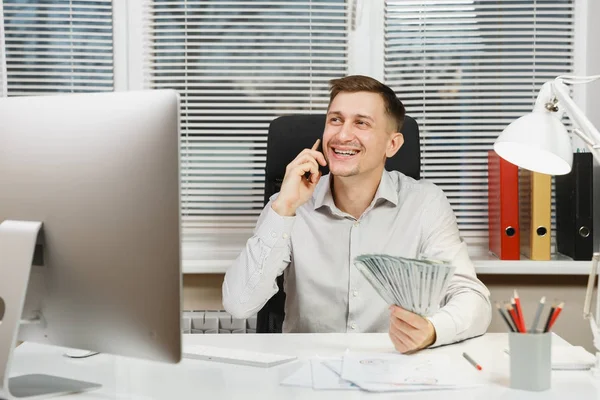 Hombre de negocios guapo sonriente en camisa sentado en el escritorio hablando en el teléfono móvil con mucho dinero en efectivo, trabajando en la computadora con monitor moderno, lámpara, documentos en la oficina de luz. Gerente o trabajador . —  Fotos de Stock