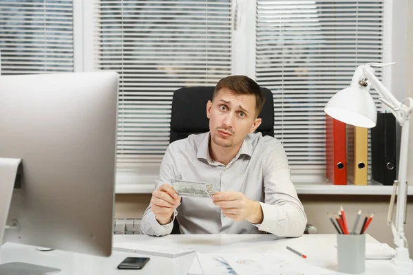 Impresionado y perplejo hombre de negocios en camisa sentado en el escritorio con mucho dinero en efectivo, trabajando en la computadora con monitor moderno, lámpara, documentos en la oficina de luz sobre el fondo de la ventana. Gerente o trabajador —  Fotos de Stock