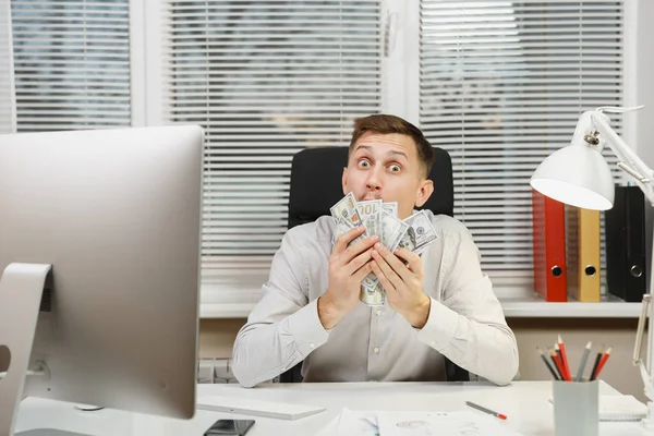 Ευτυχισμένος χαμογελαστός άνθρωπος των επιχειρήσεων στο πουκάμισο που κάθεται στο γραφείο του με πολλά μετρητά χρήματα, εργάζονται σε υπολογιστή με σύγχρονη οθόνη, εγγράφων του office φως στο φόντο του παραθύρου. Διαχείριση ή εργαζόμενος νικητής. — Φωτογραφία Αρχείου