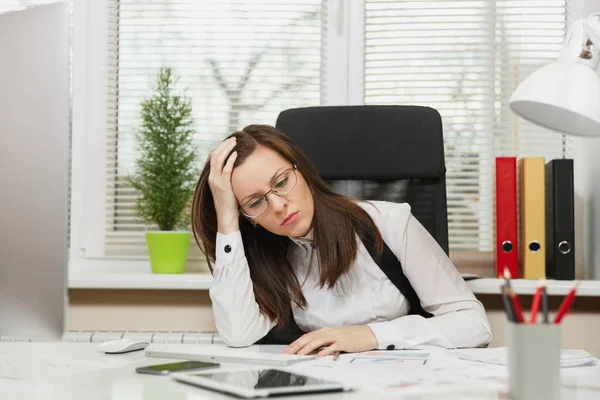 Ziemlich müde perplex und Stress braun haarige Geschäftsfrau in Anzug und Brille sitzt am Schreibtisch mit weißer Lampe, Arbeit am zeitgenössischen Computer mit Dokumenten im hellen Büro, Fensterhintergrund — Stockfoto