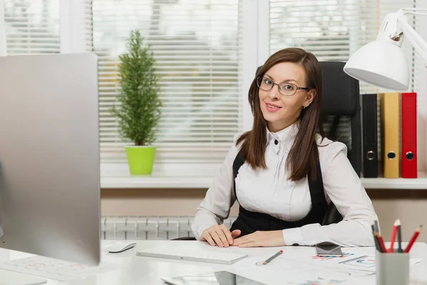 Beautiful smiling brown-hair business woman in suit and glasses sitting at the desk with tablet, working at computer with modern monitor with documents in light office, looking at the camera — Stock Photo, Image