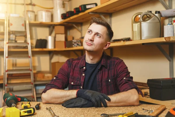 Yakışıklı beyaz genç adam ekose gömlek, siyah T-shirt, gülümseyen ahşap masa doğrama atölyesinde çalışan eldiven ile ahşap, farklı araçlar parça yer.