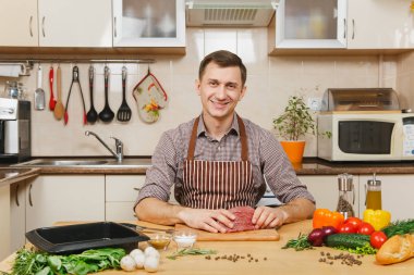 Domuz, sığır eti ya da kuzu eti hissesini ışık mutfak ahşap yüzeyli süslü mutfak eşyaları tam hazırlama evde yemek çekici beyaz delikanlı sebzelerle, masada oturan önlük.