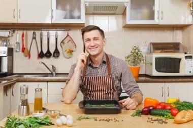 Sığır eti ya da kuzu eti hissesini ışık mutfak ahşap yüzeyli süslü mutfak eşyaları tam hazırlama evde yemek üstünde hareket eden telefon, konuşan genç adam ile sebze, masada oturan önlük.
