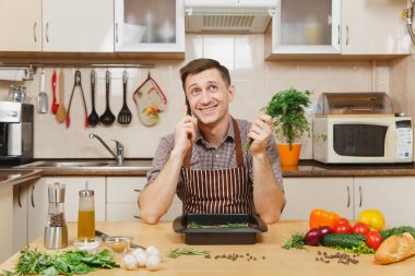Önlük ile sebze, masada oturan adam dereotu, cep telefonu, ev hazırlama et söz konusu sığır eti ya da kuzu, ahşap yüzeyli süslü mutfak eşyaları tam ışık mutfakta yemek konuşuyor.