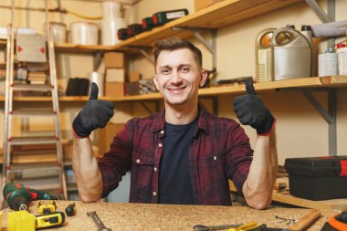 Gülümseyen beyaz yakışıklı genç başparmak yukarı, ahşap masa yer ahşap, farklı araçları parçası ile doğrama atölyesinde çalışan gösterilen ekose gömlek, siyah T-shirt, eldiven.