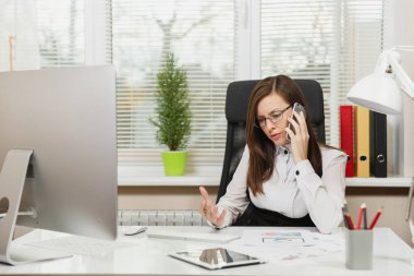 Işık Office belgeleri ile çağdaş bilgisayarda çalışan güzel ciddi iş kadın takım elbise ve masa başında oturan gözlük cep telefonu üzerinde sorunları çözme bir kenara seyir konuşurken