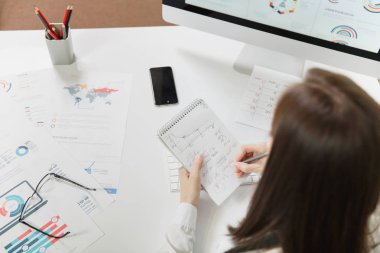 İş kadını masada oturan takım elbise, dizüstü bilgisayar, cep telefonu ve klavye arka plan üzerinde ışık Office belgeleri ile bilgisayarda çalışan için bilgileri kalem ile yazma, arkadan görünüm