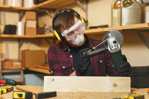 Caucasian young man in plaid shirt, black T-shirt, noise insulated headphones, protective mask working in carpentry workshop at wooden table place with different tools, sawing wood with power saw. — Stock Photo, Image