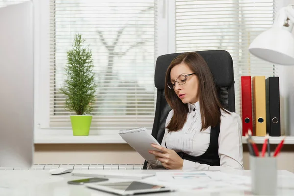 Beautiful serious and engrossed brown-hair business woman in suit and glasses sitting at the desk with tablet, working at computer with modern monitor with documents in light office, looking aside — Stock Photo, Image