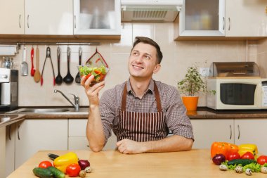 Gülümseyen Avrupa yakışıklı genç hafif modern mutfak kase sebze salatası ile masada oturan önlük, kahverengi gömlekli. Diyet kavramı. Sağlıklı bir yaşam tarzı. Evde yemek. Yemek hazırlamak.