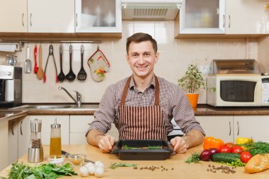 Domuz, sığır eti ya da kuzu eti hissesini ışık mutfak ahşap yüzeyli süslü mutfak eşyaları tam hazırlama evde yemek beyaz yakışıklı genç ile sebze, masada oturan önlük.