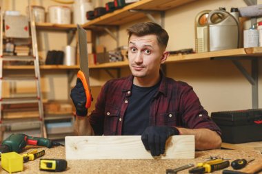 Yakışıklı karizmatik gülümseyen beyaz genç adam ekose gömlek, siyah T-shirt, testere, ahşap masa doğrama atölyesinde çalışan ile ahşap testere eldiven ile ahşap, farklı araçlar parça yerleştirin..