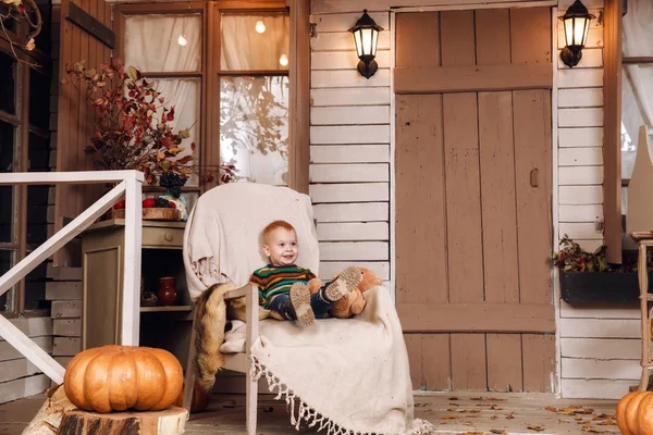 Χαριτωμένο μικρό αγόρι μωρό ντυμένος με ένα πουλόβερ, τζιν που κάθεται σε καρέκλα κοντά στο σπίτι με λούτρινο αρκουδάκι στο φθινόπωρο. Αφήνει στην αυλή, φωτίζονται από ΦΑΚΟΙ, με ξηρά πτώση κίτρινο, πορτοκαλί κολοκύθες. — Φωτογραφία Αρχείου