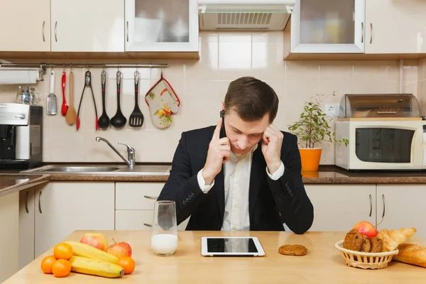 Κουρασμένος λυπημένος νέοι επιχείρηση άνθρωπος στο κοστούμι, πουκάμισο, έχοντας πρωινό, κάθεται στο τραπέζι με το tablet, μιλώντας σε κινητό τηλέφωνο, τρώγοντας φρούτα στην ελαφριά κουζίνα. Υγιεινού τρόπου ζωής. Το μαγείρεμα στο σπίτι. Προετοιμασία φαγητού. — Φωτογραφία Αρχείου