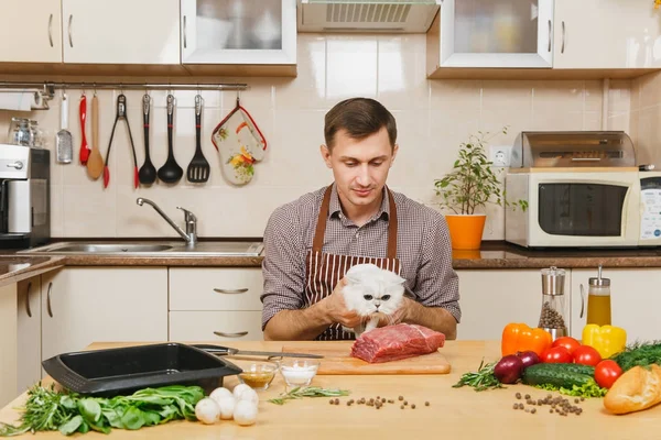 stock image Young man in apron sitting with furry cat at table with vegetables, cooking at home preparing meat whitestake from pork, beef or lamb, in light kitchen with wooden surface, full of fancy kitchenware.