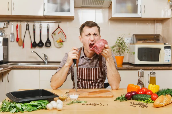 Привлекательный кавказский молодой человек в фартуке сидит за столом с овощами, готовит дома мясной кол из свинины, говядины или баранины, на светлой кухне с деревянной поверхностью, полной модной кухонной утвари . — стоковое фото