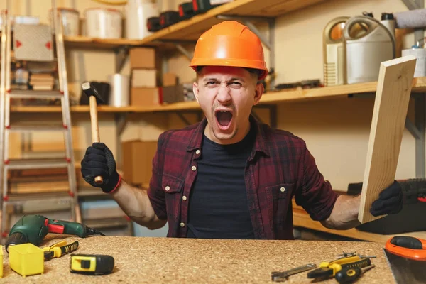 Crazy agressif jeune homme caucasien en colère en chemise à carreaux, T-shirt noir, casque de protection orange, gants travaillant dans l'atelier de menuiserie à la place de la table en bois avec un morceau de marteau de bois différents outils — Photo