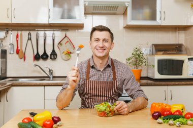 Işık mutfakta yemek çekici gülümseyen Beyaz delikanlı kase sebze salatası ile masada oturan önlük, kahverengi gömlekli. Diyet kavramı. Sağlıklı bir yaşam tarzı. Evde yemek. Yemek hazırlamak.