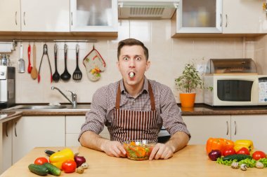 Kase ve Bıldırcın yumurta içinde ağız açık mutfakta sebze salatası ile masada oturan önlük, kahverengi gömlekli eğlenceli beyaz genç adam. Diyet kavramı. Sağlıklı bir yaşam tarzı. Evde yemek. Yemek hazırlamak.