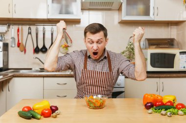 Çatal ve bıçak ışık mutfakta sebze salatası saldıran öfkeli deli aç genç adam masadan, önlük, kahverengi gömlekli. Diyet kavramı. Sağlıklı bir yaşam tarzı. Evde yemek. Yemek hazırlamak.