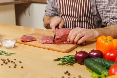 Domuz, sığır eti ya da kuzu eti hissesini ışık mutfak ahşap yüzeyli süslü mutfak eşyaları tam hazırlama evde yemek masada oturan sebze ile önlük beyaz genç adam kadar kapatın.