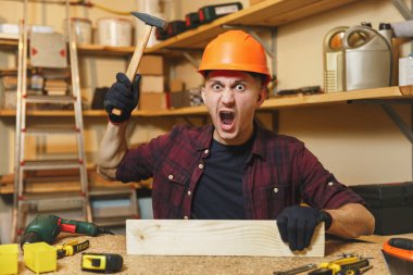Çılgın agresif kızgın beyaz genç adam ekose gömlek, siyah T-shirt, turuncu koruyucu kask, eldiven ahşap masa doğrama atölyesinde çalışan tahta çekiç farklı araçlar parça ile yerleştirin.