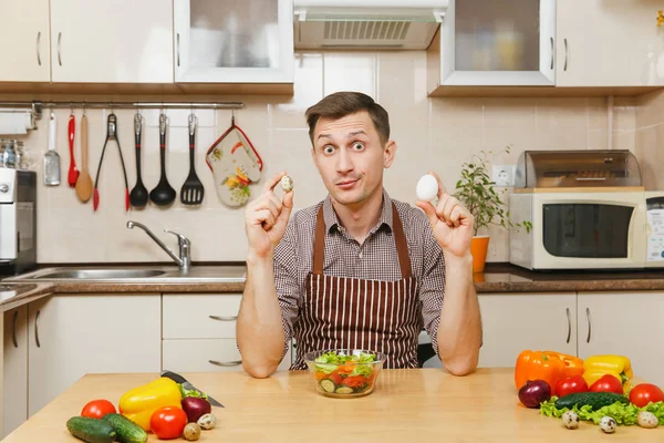 Schöner kaukasischer junger Mann in Schürze, braunes Hemd am Tisch sitzend, Hühner- und Wachteleier in der Hand, Gemüsesalat in heller Küche. Diätkonzept. Gesunder Lebensstil. Kochen zu Hause. Essen zubereiten — Stockfoto