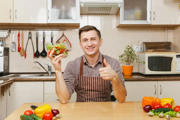 Jolie jeune homme caucasien en tablier, chemise brune assise à table avec salade de légumes dans un bol, montrant pouce vers le haut dans la cuisine légère. Concept de régime. Mode de vie sain. Cuisiner à la maison. Préparer les aliments — Photo