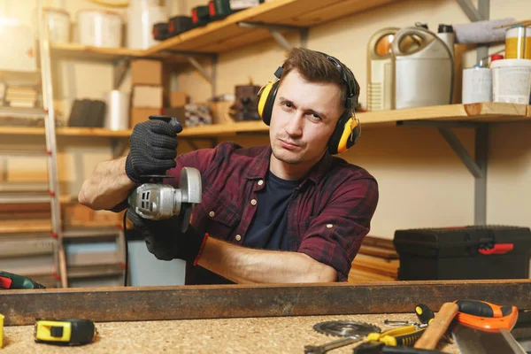 Guapo joven caucásico sonriente en camisa a cuadros, camiseta negra, auriculares con aislamiento acústico que trabajan en el taller de carpintería en el lugar de mesa de madera con diferentes herramientas, plancha de aserrar con sierra eléctrica . — Foto de Stock