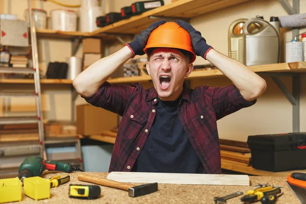 Loco agresivo enojado caucásico joven en camisa a cuadros, camiseta negra, casco protector naranja, guantes que trabajan en taller de carpintería en lugar de mesa de madera con pieza de martillo de madera diferentes herramientas — Foto de Stock