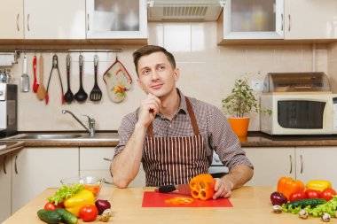 Gülümseyen beyaz yakışıklı genç apronda, kahverengi gömlek masadan, sebze salatası için ışık mutfakta bıçakla keser. Diyet kavramı. Sağlıklı bir yaşam tarzı. Evde yemek. Yemek hazırlamak.