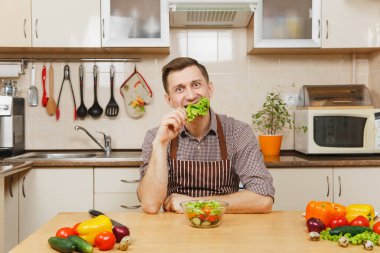 Yakışıklı beyaz delikanlı apronda, kahverengi gömlek masada oturan yetişmek ve marul sebze salatası için ışık mutfakta bıçakla yırtılma. Diyet kavramı. Evde yemek. Yemek hazırlamak.