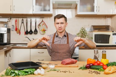 Kızgın genç adam sebze ile masada oturan önlük, evde yemek gösteren başparmak aşağı, sığır eti veya domuz eti hissesini ahşap yüzeyli açık mutfak hazırlanıyor süslü mutfak eşyaları tam.