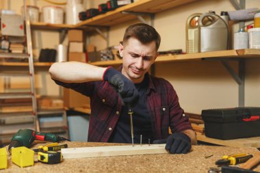 Yakışıklı gülümseyen beyaz genç adam ekose gömlek, siyah T-shirt, tornavida vida, ahşap masa doğrama atölyesinde çalışan tarafından büküm eldiven ile ahşap, farklı araçlar parça yerleştirin..