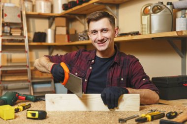 Yakışıklı karizmatik gülümseyen beyaz genç adam ekose gömlek, siyah T-shirt, testere, ahşap masa doğrama atölyesinde çalışan ile ahşap testere eldiven ile ahşap, farklı araçlar parça yerleştirin..