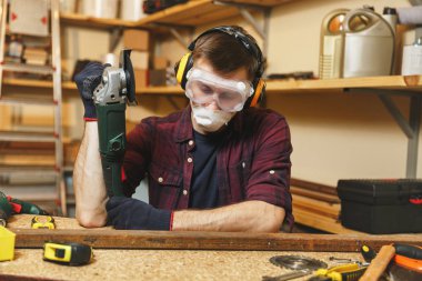 Ekose gömlek, siyah T-shirt, ses yalıtımlı kulaklık beyaz genç adam, koruyucu maske ahşap masa yer farklı araçlar, güç ile Demir testere ile doğrama atölyesinde çalışan gördüm.