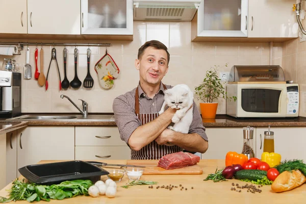 Young man in apron sitting with furry cat at table with vegetables, cooking at home preparing meat whitestake from pork, beef or lamb, in light kitchen with wooden surface, full of fancy kitchenware.